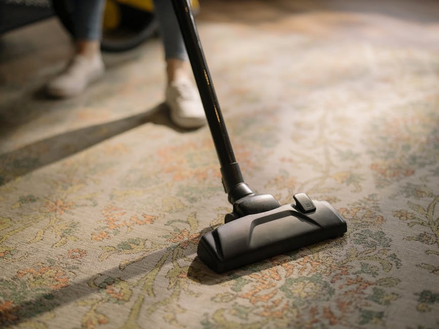 A close-up view of a vacuum cleaner head being used on a patterned residential carpet, showing a combination of floral and geometric designs in soft pastel shades. The vacuum cleaner is black and grey