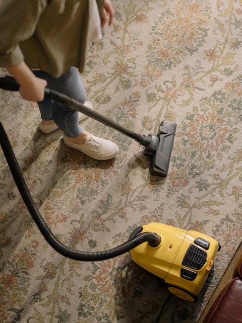 A close-up view of a vacuum cleaner head being used on a patterned residential carpet, showing a combination of floral and geometric designs in soft pastel shades. The vacuum cleaner is black and grey