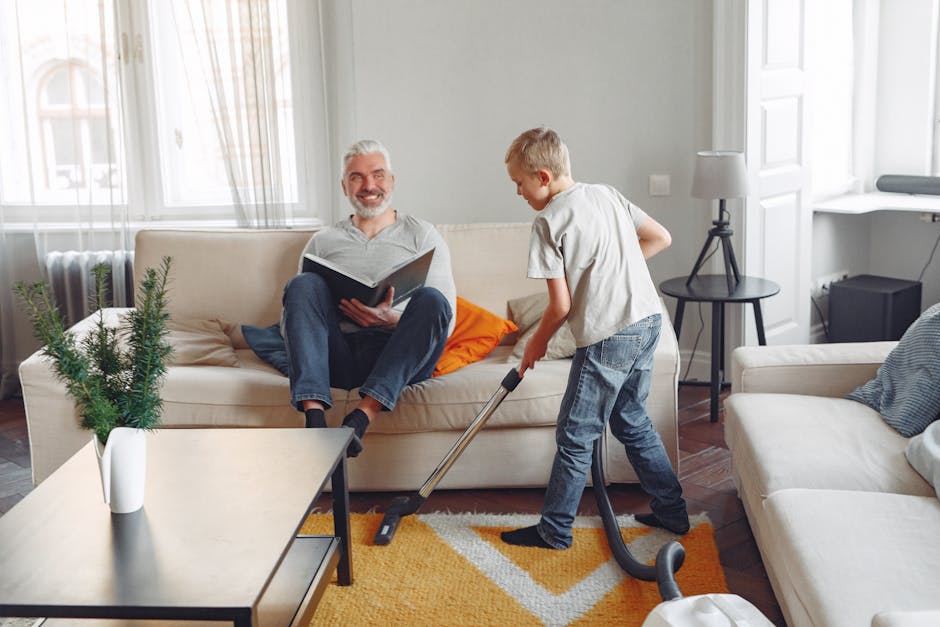A yellow cylindrical vacuum cleaner with a black hose attachment is placed on a patterned area rug in a living room. The room features wooden flooring, a low black metal console table against the whit